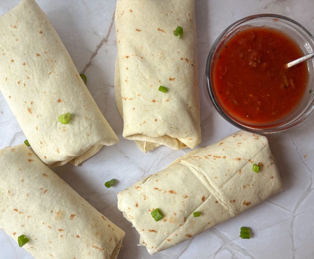 Four breakfast burritos without eggs laid out on a table with a side of salsa next to it.