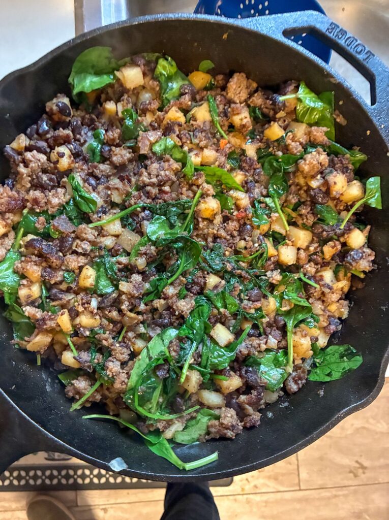 Breakfast potatoes, spices, spinach, and black beans in a large cast iron skillet
