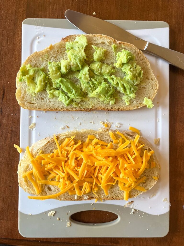 2 slices of sourdough bread. One covered with mashed avocado and one covered with shredded cheddar cheese.