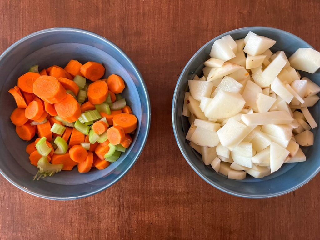 A bowl full of chopped carrots and celery and another bowl full of peeled and chopped potatoes