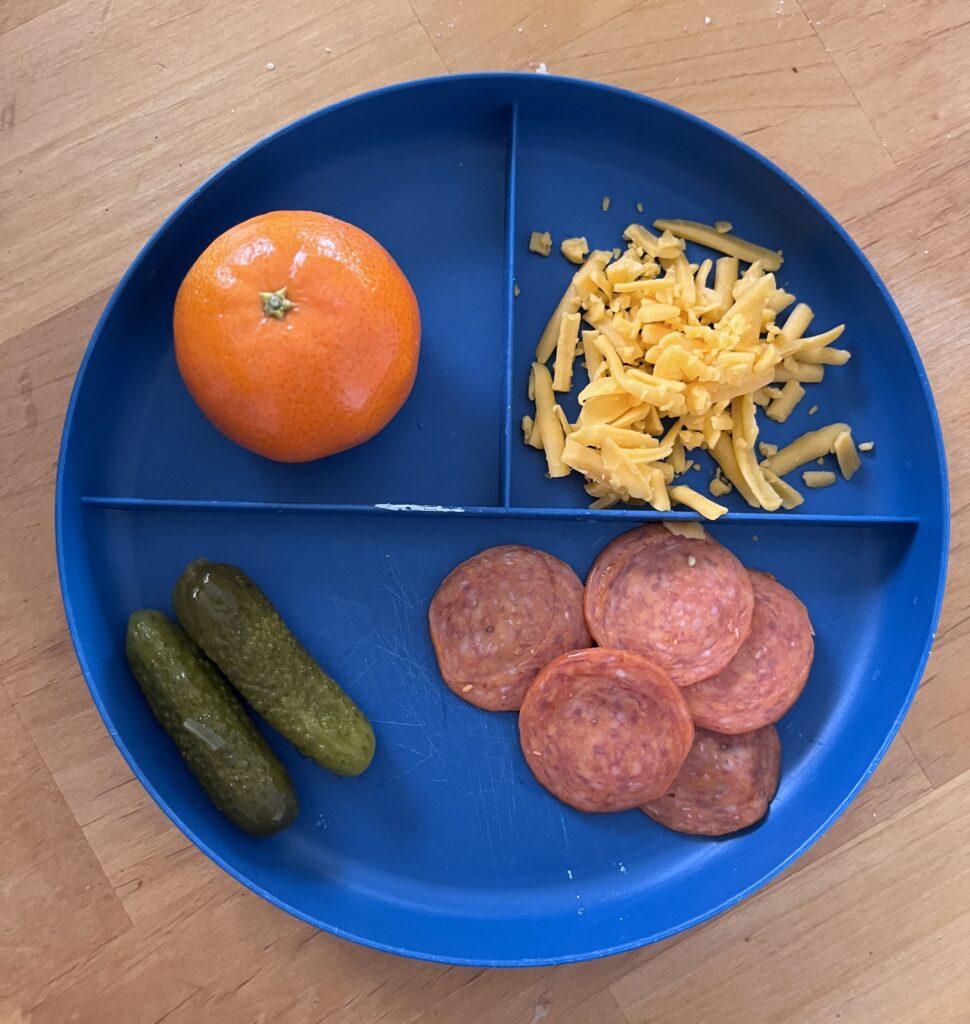 Easy preschool lunch on plate including pepperoni, clementine, shredded cheese, and pickles