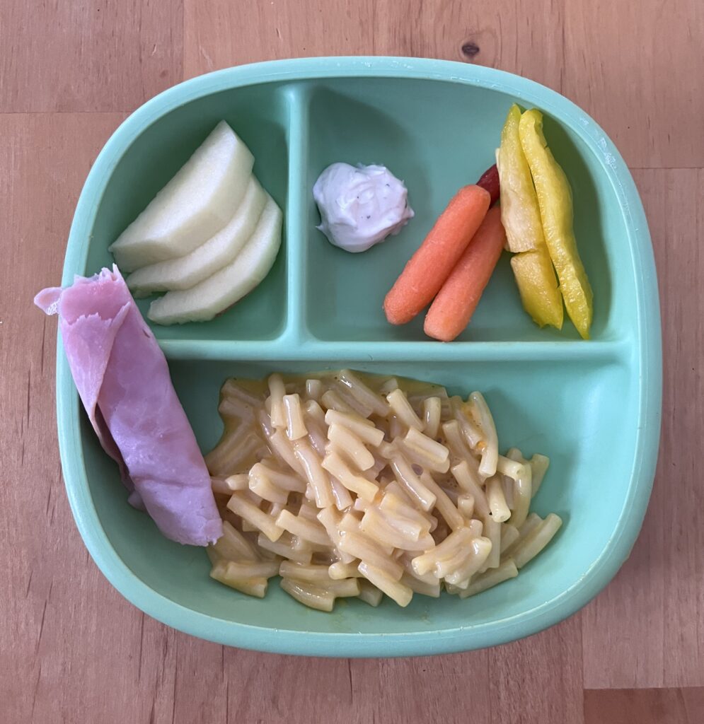 Easy toddler lunch on a plate including macaroni and cheese, sliced deli ham, vegetables and dip, and sliced apples