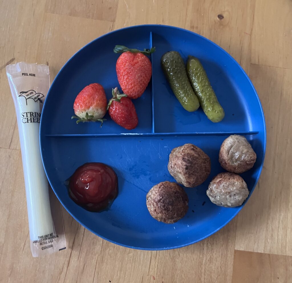 Easy preschool lunch ideas on a plate including strawberries, pickles, cheese stick, and meatballs