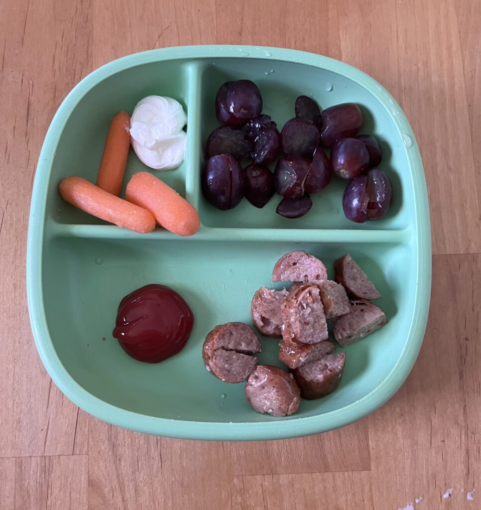 Preschool lunch on a plate of chicken sausage, ketchup, grapes, and carrots and ranch dip