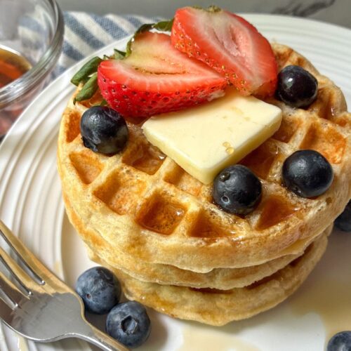 Eggless waffles stacked on top of each other with fresh fruit and a fork on a plate
