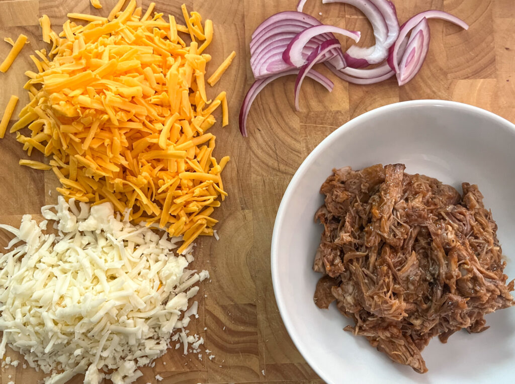 Ingredients in BBQ pulled pork pizza laid out on a cutting board including cheese, onion, and pulled pork