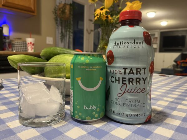 A small glass filled with ice next to a lime bubbly and a jar of tart cherry juice