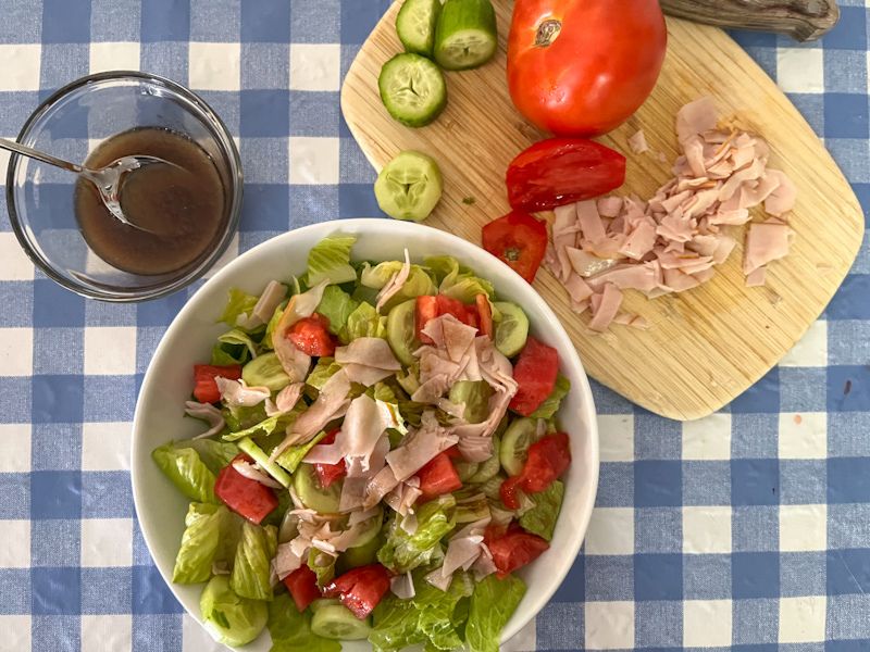 Bowl of salad and a bowl of salad dressing sitting next to it. A cutting board with vegetables and meat on it for the salad.