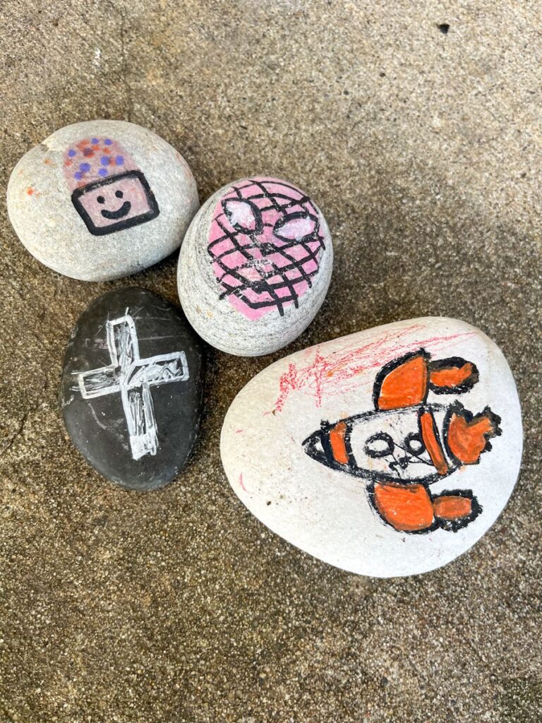 4 painted rocks with images of a cross, cupcake, spiderman, and rocket.