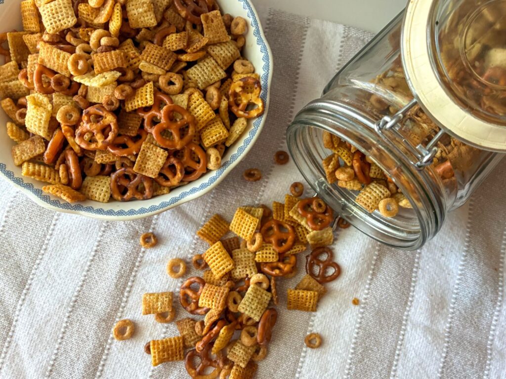 Oven baked chex mix spilling out of a container with a bowl full of the Chex mix sitting next to it.