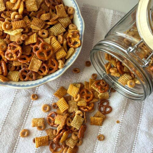 Oven baked chex mix spilling out of a container with a bowl full of the Chex mix sitting next to it.