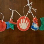5 salt dough ornaments in a row that are painted. A handprint ornament, star, snowman, and stocking