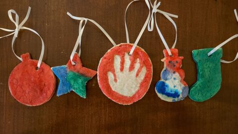 5 salt dough ornaments in a row that are painted. A handprint ornament, star, snowman, and stocking