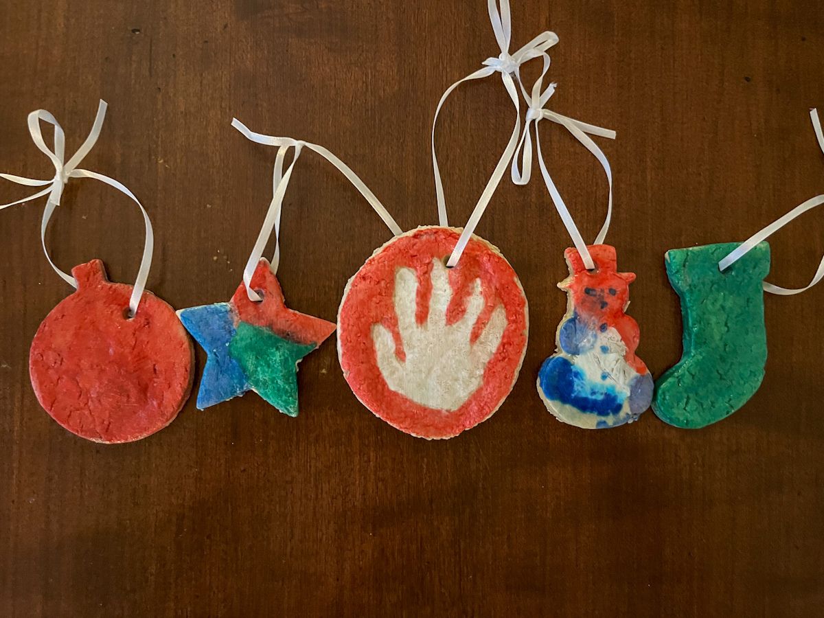 5 salt dough ornaments in a row that are painted. A handprint ornament, star, snowman, and stocking