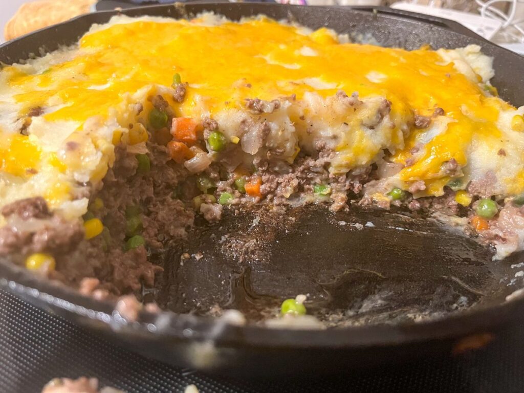 shepherds pie in a cast iron skillet with some scooped out so you can see the layers of meat, veggies, and potatoes.