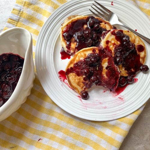 3 pancakes covered in blueberry maple syrup and a dish filled with the syrup