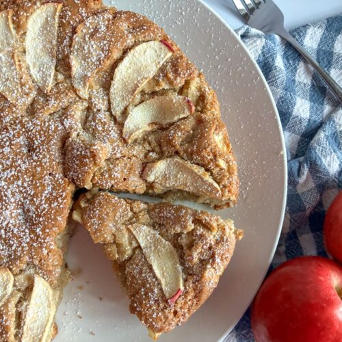 Eggless apple cake served on a plate with one slice cut out. 2 apples on the side of the plate.
