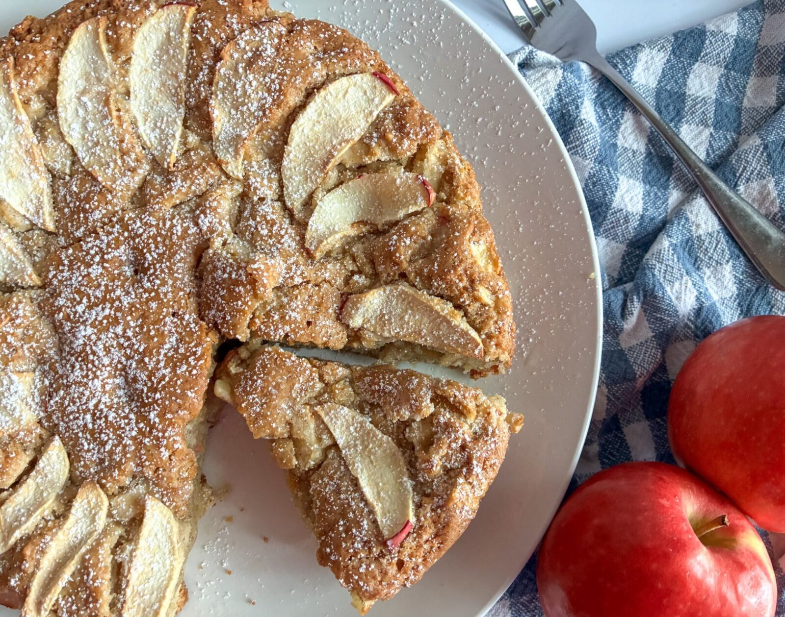 Eggless apple cake served on a plate with one slice cut out. 2 apples on the side of the plate.