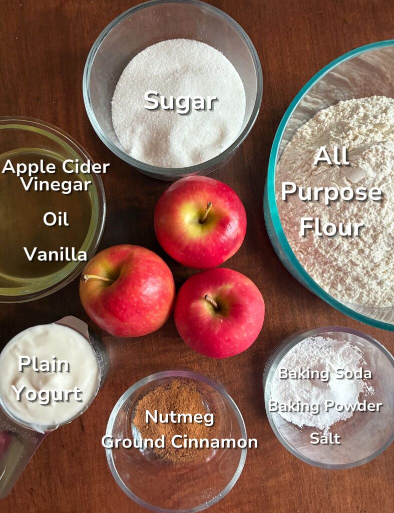 All ingredients laid out on a table for eggless apple cake. Text label on top of the ingredients.