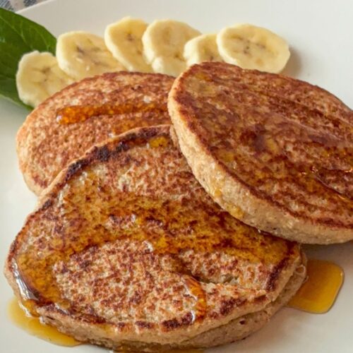 Three oatmeal blender pancakes on a plate with sliced bananas behind them.