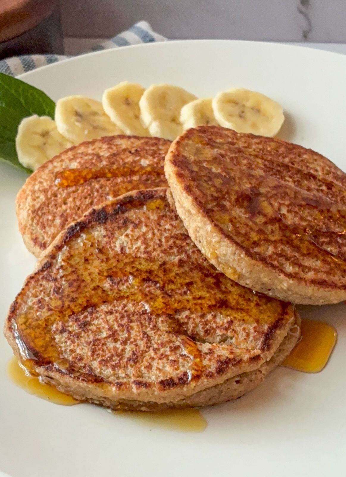 Three oatmeal blender pancakes on a plate with sliced bananas behind them.