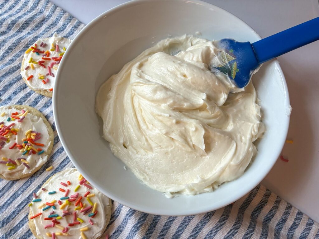 Large bowl of icing being scooped out with a spatula with cookies laying on the side.