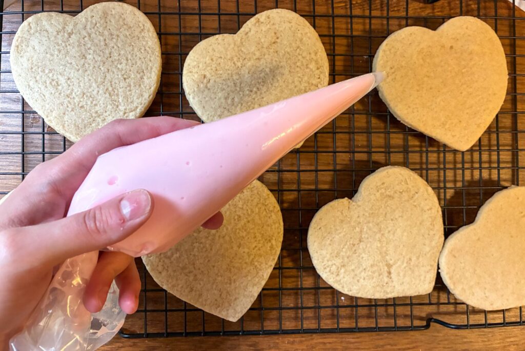Hand holding a piping bag with eggless royal icing above heart shaped sugar cookies