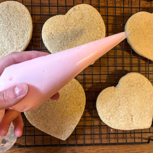 Hand holding a piping bag with eggless royal icing above heart shaped sugar cookies