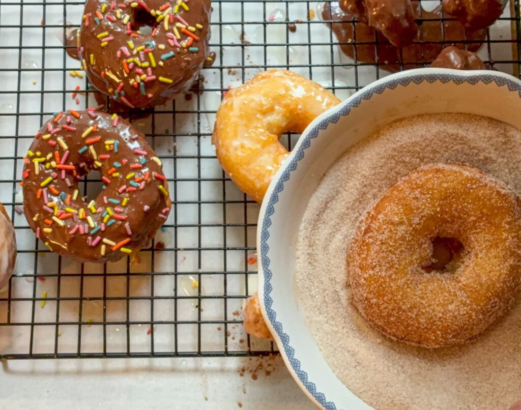 Multiple donuts on a cooling rack and a cinnamon sugar donut sitting in a bowl being dipped