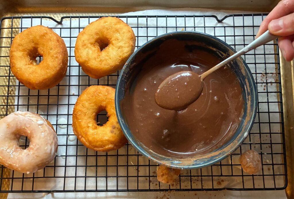 Four donuts on a cooling rack with a small bowl with chocolate glaze in it